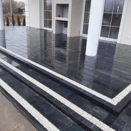 Firma Brukpat Radosław Mężyk - Nowoczesny taras z ciemnej kostki brukowej z białymi akcentami na schodach, widok na kolumny podtrzymujące zadaszenie i murowany grill.