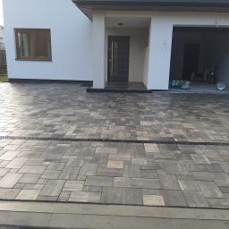 Firma Brukpat Radosław Mężyk - Ułożona szara kostka brukowa na podjeździe przed nowoczesnym domem z białymi ścianami, widoczny odpływ liniowy.