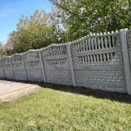 Długi, prefabrykowany, betonowy płot w odcieniach szarości, z dekoracyjnymi elementami i wzorem imitującym cegłę, oddzielający trawnik od chodnika, na tle zieleni drzew i błękitnego nieba.