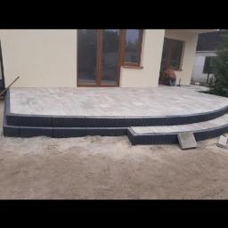 Firma Brukpat Radosław Mężyk - Wykończony taras z jasnoszarej kostki brukowej przed beżowym domem z brązowymi oknami, widoczny pracownik w trakcie fugowania.