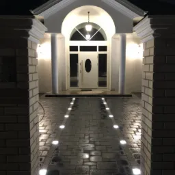Eleganckie wejście do domu z ciemnoszarej kostki z wbudowanymi lampami punktowymi, oświetlające białe kolumny i łuk nad drzwiami wejściowymi pod wieczornym niebem.