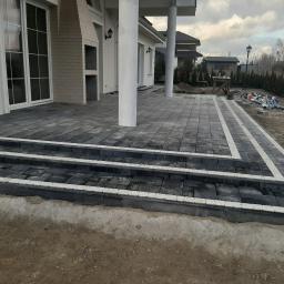 Firma Brukpat Radosław Mężyk - Nowoczesny taras z ciemnej kostki brukowej z jasnymi obrzeżami, schody prowadzące do budynku, w tle pracownik w kombinezonie pomarańczowym wykonujący prace wykończeniowe.