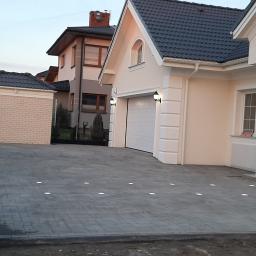 Firma Brukpat Radosław Mężyk - Szara kostka brukowa na podjeździe do domu z oświetleniem wbudowanym w nawierzchnię, widok na dom z garażem i fragment posesji sąsiada.