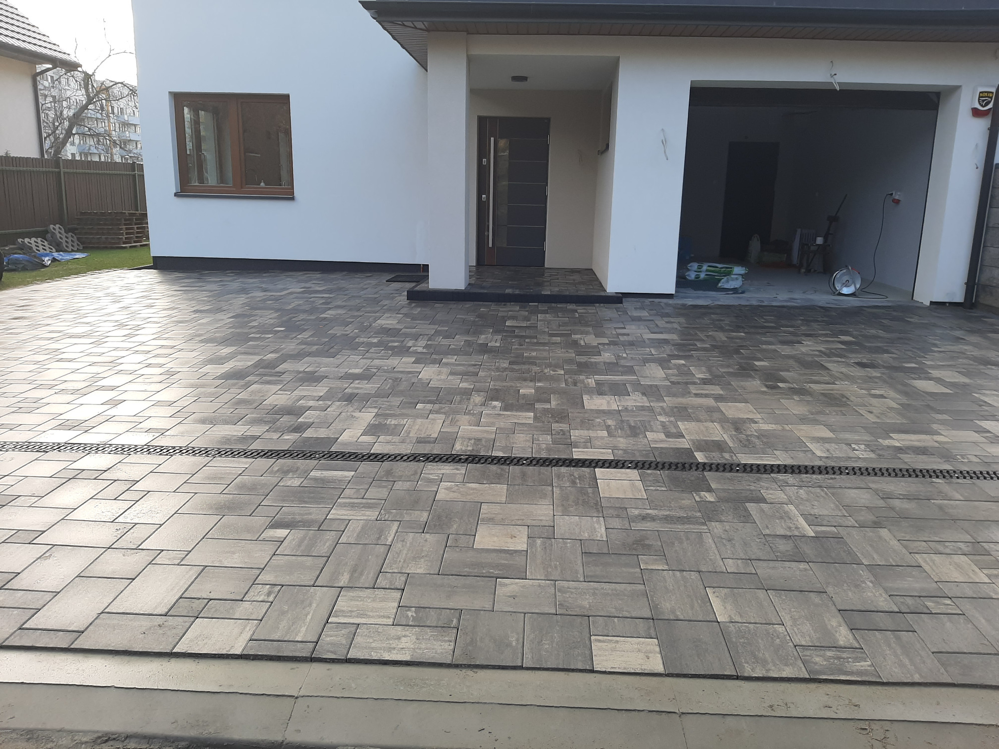 Ułożona szara kostka brukowa na podjeździe przed nowoczesnym domem z białymi ścianami, widoczny odpływ liniowy.