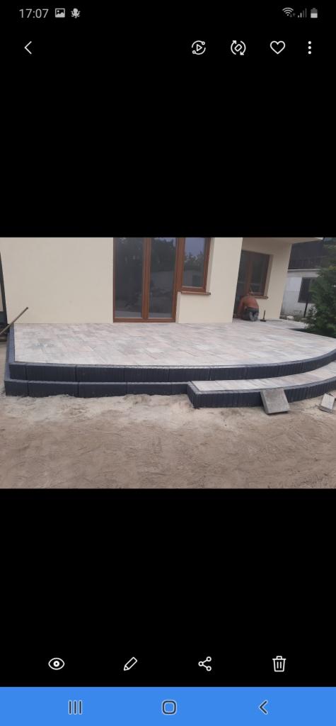 Wykończony taras z jasnoszarej kostki brukowej przed beżowym domem z brązowymi oknami, widoczny pracownik w trakcie fugowania.