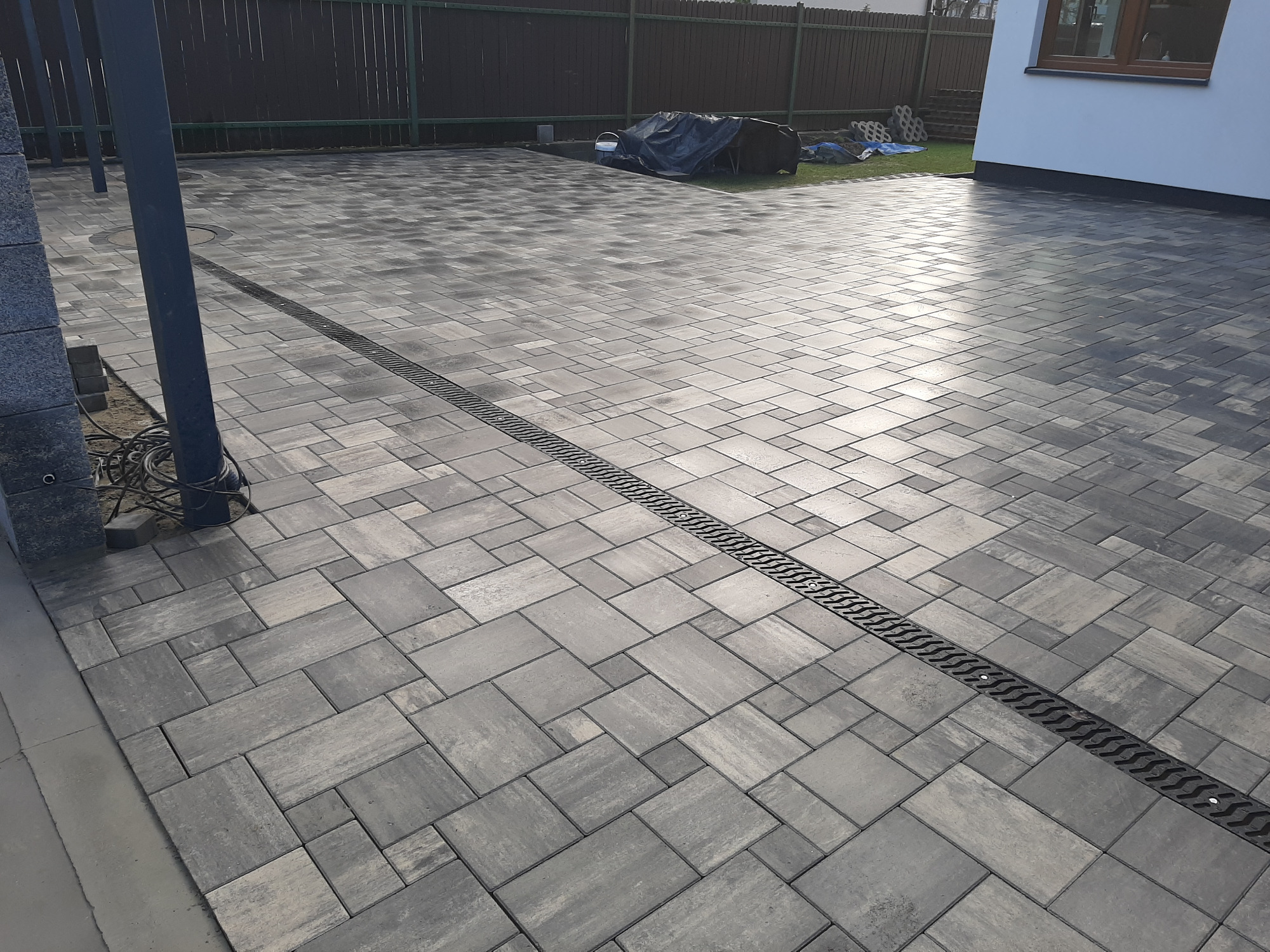 Ułożona szara kostka brukowa z odpływem liniowym na posesji, widoczny fragment domu z oknem i ogrodzenie w tle.