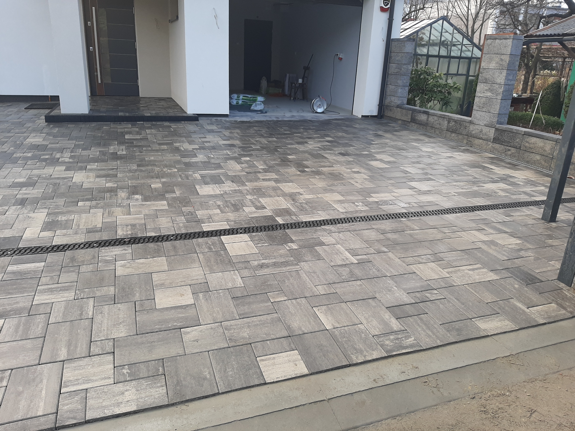 Szara, ułożona kostka brukowa przed nowoczesnym domem z otwartym garażem, widoczny system odwadniający liniowy i fragment szklarni w tle.