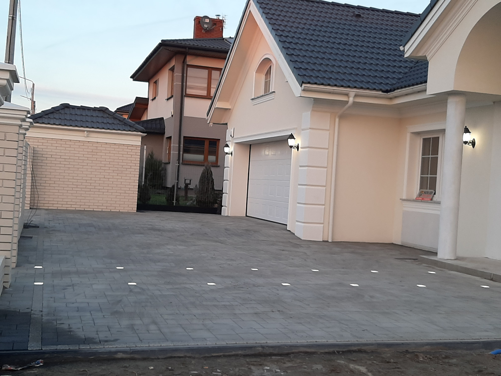 Szara kostka brukowa na podjeździe do domu z oświetleniem wbudowanym w nawierzchnię, widok na dom z garażem i fragment posesji sąsiada.