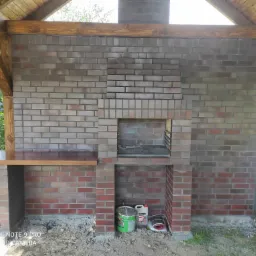 Murowana konstrukcja grilla z cegły klinkierowej pod drewnianą wiatą, z blatem roboczym z drewna, widoczne narzędzia i materiały budowlane w dolnej części.