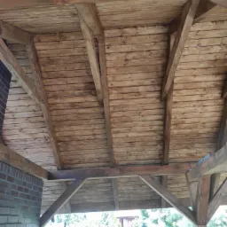 Drewniana konstrukcja dachu altany z widocznymi belkami i deskami, pomalowana bejcą na ciepły brązowy kolor, z fragmentem murowanej ściany i zielenią w tle.