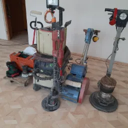 Parkiet ułożony w geometryczny wzór, z widocznymi maszynami do cyklinowania i szlifowania podłóg, w tym maszyna marki Flex z oświetleniem, oraz odkurzacz przemysłowy Festool.