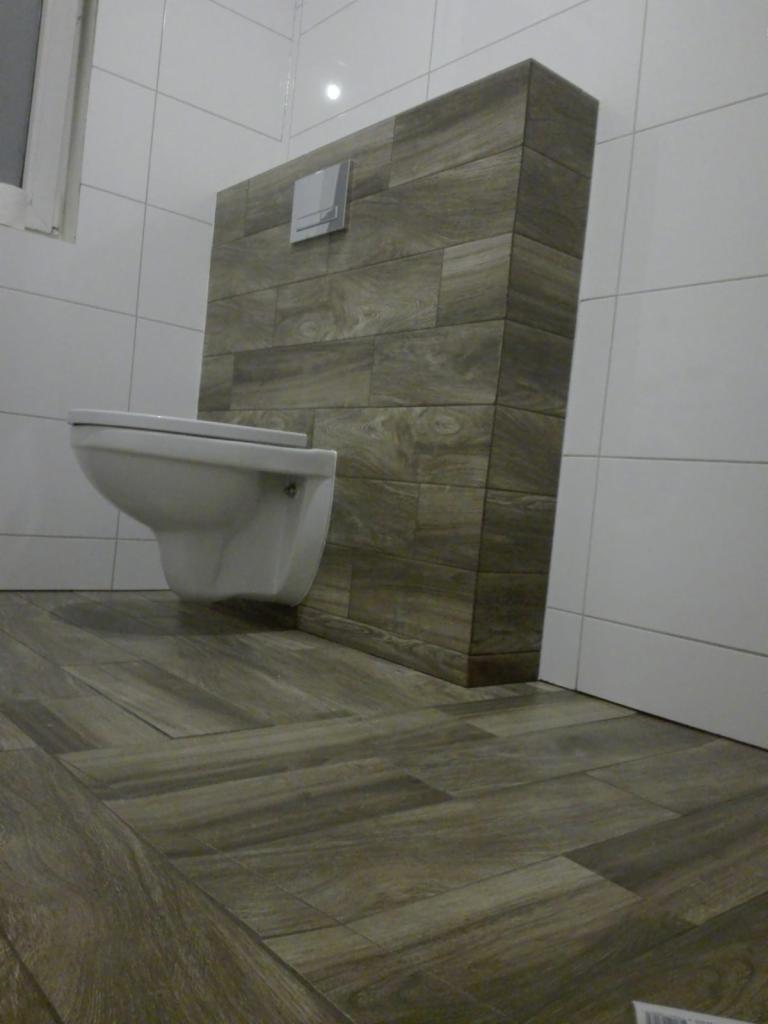 Nowoczesna toaleta wisząca z zabudową z płytek imitujących drewno, dopasowanych do podłogi, białe płytki na ścianach, minimalistyczny design.