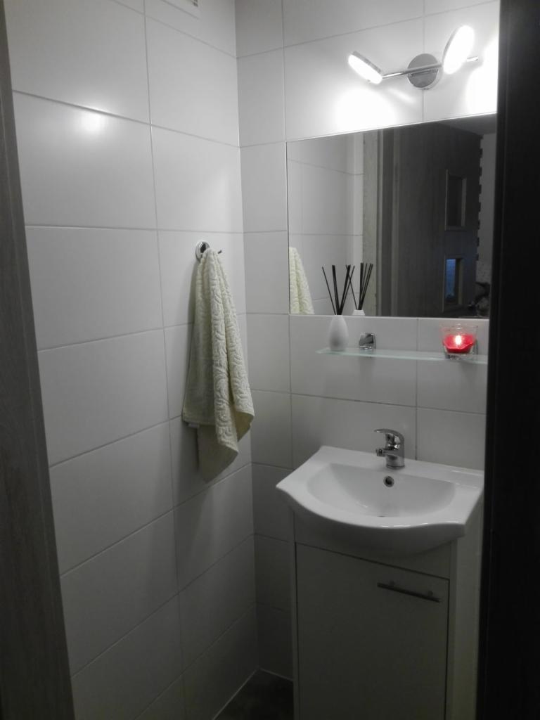 Biała łazienka z umywalką, szafką pod umywalką, lustrem, ręcznikiem na wieszaku i białymi płytkami na ścianach.