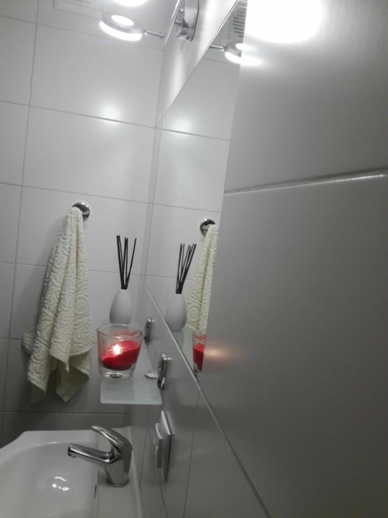 Biała łazienka z umywalką, baterią, lustrem, ręcznikiem, świecą w szklanym naczyniu i dyfuzorem zapachowym na szklanej półce.