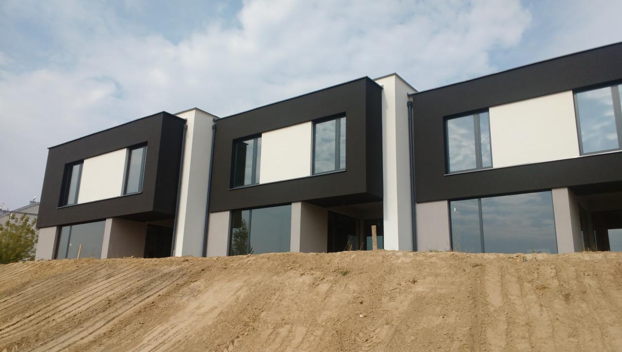 Trzy segmenty nowoczesnego domu w trakcie budowy, z czarnymi i białymi panelami elewacyjnymi, widoczne duże okna i nasyp ziemi przed budynkiem.