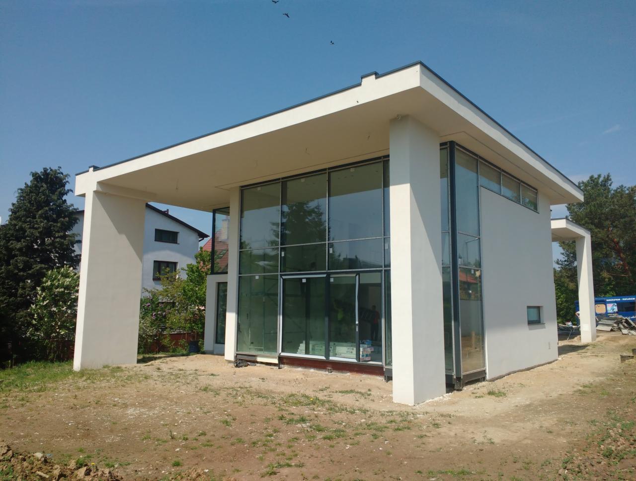 Nowoczesny budynek z dużymi przeszkleniami i białymi elementami konstrukcyjnymi, otoczony niezagospodarowanym terenem, widoczne odbicia w szybach.