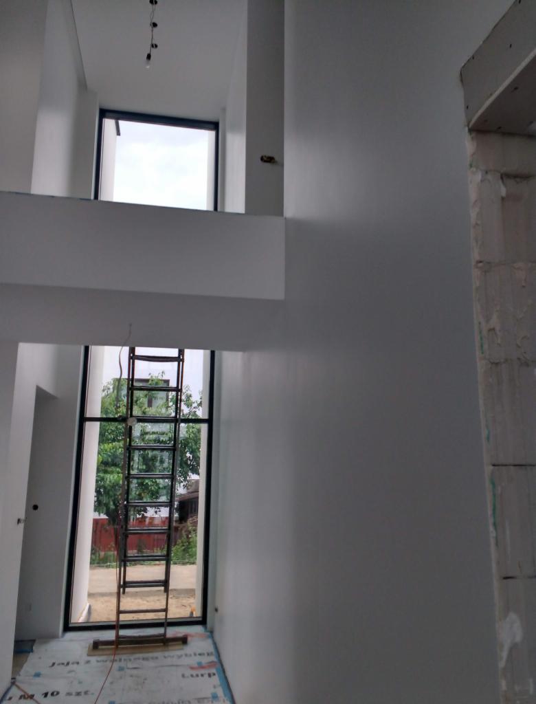 Wnętrze dwupoziomowego pomieszczenia po tynkowaniu maszynowym, widoczna drabina przy oknie i surowy fragment ściany z prawej strony.