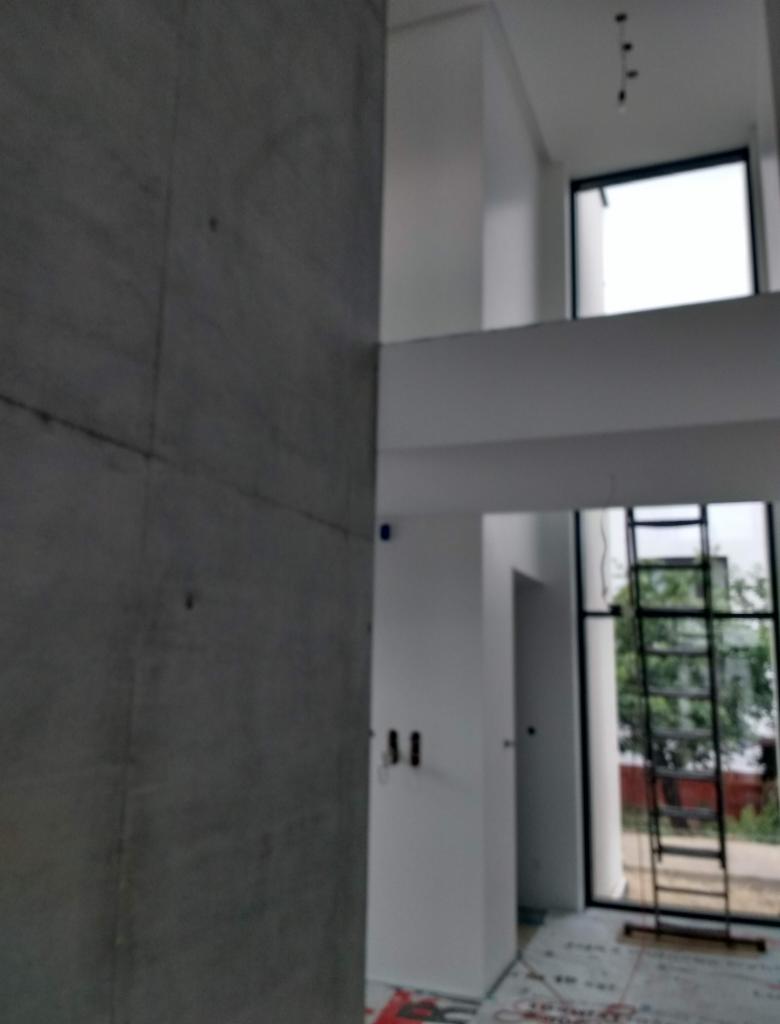Wnętrze w trakcie wykańczania: surowa ściana z betonu architektonicznego kontrastuje z pomalowanymi na biało ścianami, widoczna drabina oparta o okno oraz zabezpieczona podłoga.