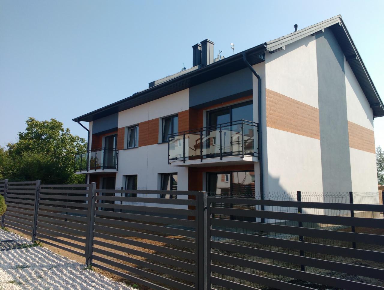 Dwukondygnacyjny budynek mieszkalny z balkonami, wykończony w odcieniach bieli, szarości i drewna, ogrodzony metalowym płotem na tle błękitnego nieba.