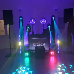 Scena DJ'a Oscar oświetlona dynamicznymi światłami w kolorach niebieskim, zielonym i różowym. Widoczne głośniki, reflektory i panel DJ'ski z logo.