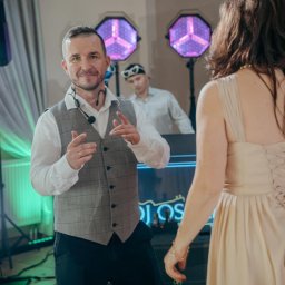 Oskar Wawrzynowicz - Uśmiechnięty wodzirej w kamizelce gestykuluje na parkiecie, w tle DJ z okularami w kształcie serc i efekty świetlne podczas imprezy.