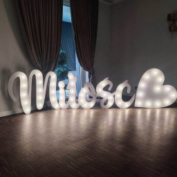 Oskar Wawrzynowicz - Świecący napis 'Miłość' z żarówkami na drewnianej podłodze, ustawiony przed oknem z zasłonami. Dekoracja na imprezę, romantyczny nastrój.