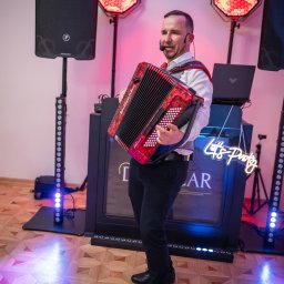 Oskar Wawrzynowicz - Akordeonista w trakcie występu, z mikrofonem nagłownym, na tle oświetlenia scenicznego i neonu 'Let's Party'. Widoczny parkiet i sprzęt nagłośnieniowy.