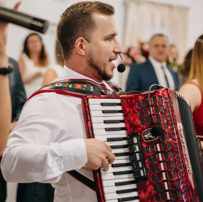 Akordeonista w białej koszuli gra na czerwonym akordeonie na tle rozmytej publiczności podczas występu na imprezie.