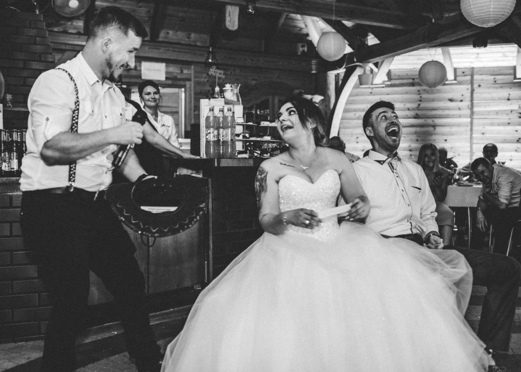 Czarno-białe zdjęcie z wesela: wodzirej z mikrofonem rozbawia parę młodą siedzącą na krzesłach, w tle goście weselni i bar. Radosna atmosfera.