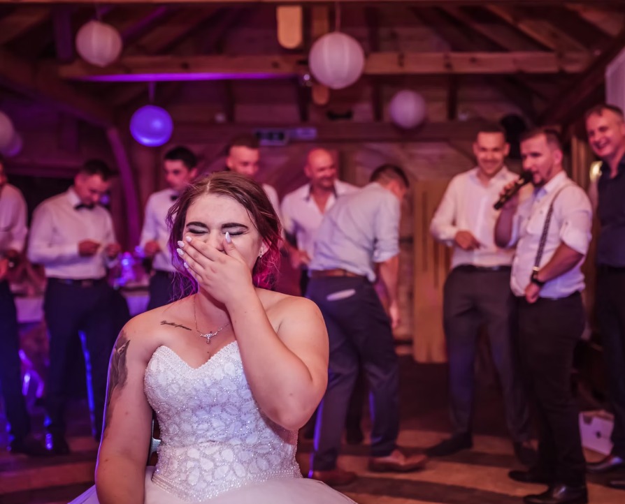 Wzruszona panna młoda w sukni ślubnej, zasłaniająca twarz dłonią, w tle muzycy z mikrofonem i goście weselni w sali z drewnianą konstrukcją i lampionami.