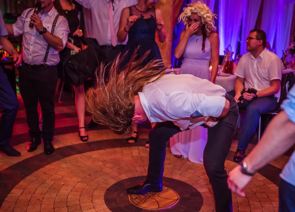 Dynamiczne ujęcie tańczącego mężczyzny z długimi włosami na parkiecie w otoczeniu innych osób podczas imprezy, w tle widoczna scena i oświetlenie.