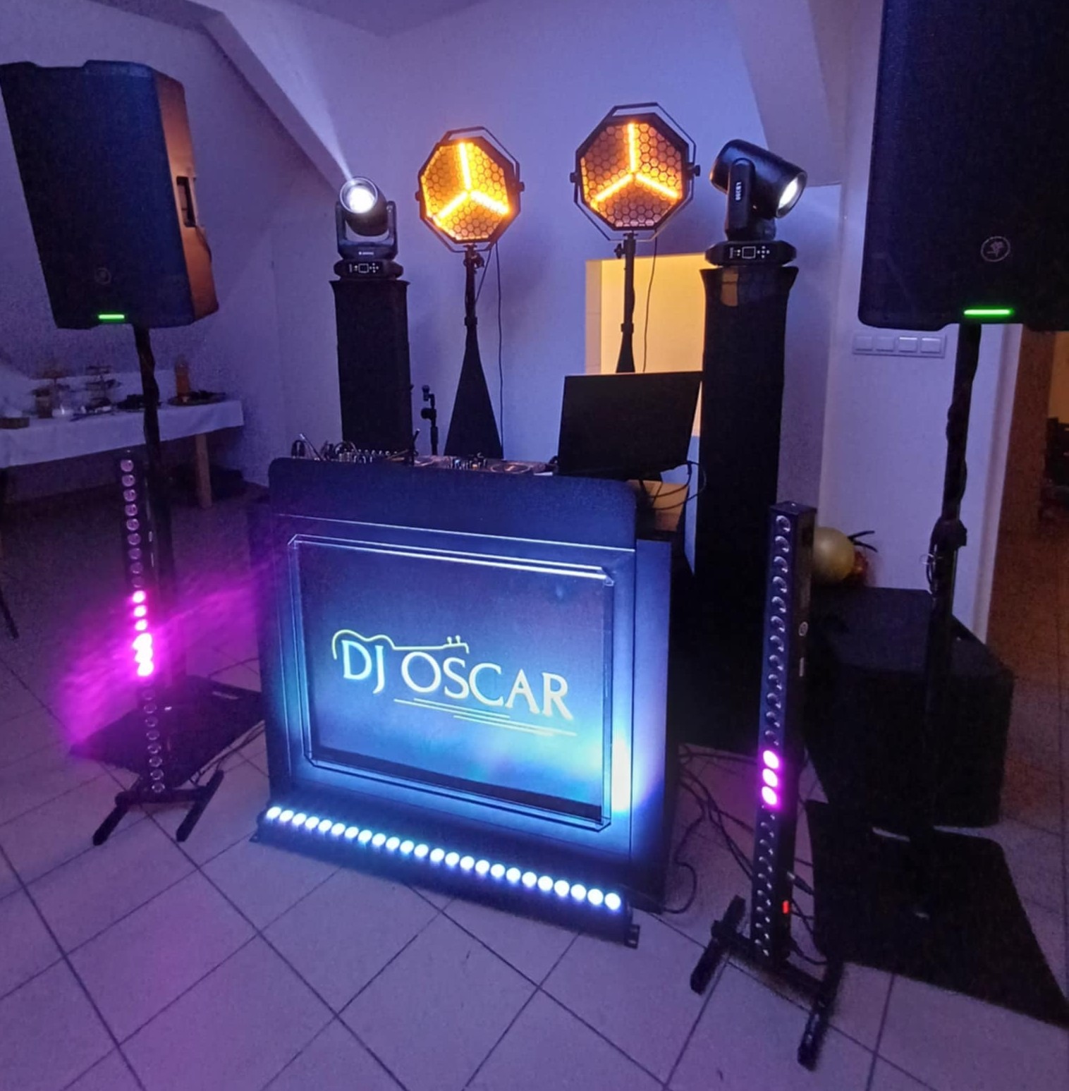 Stanowisko DJ-a z oświetleniem scenicznym, kolumnami głośnikowymi i panelem z napisem 'DJ Oscar', oświetlone fioletowymi i pomarańczowymi światłami.