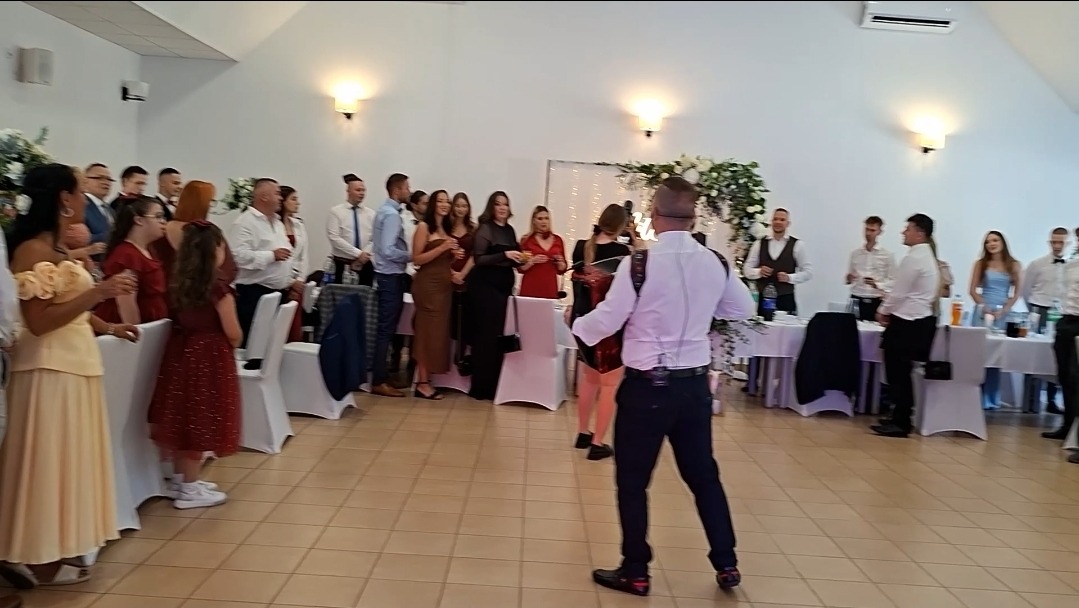 Akordeonista w akcji podczas wesela, goście weselni w tle. Sala udekorowana kwiatami, białe krzesła.