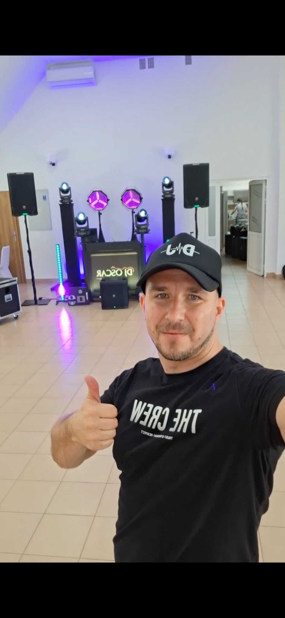DJ Oscar prezentuje sprzęt oświetleniowy i nagłośnieniowy na sali. W tle widoczna osoba. Uśmiechnięty mężczyzna w czapce z logiem DJ daje kciuk w górę.