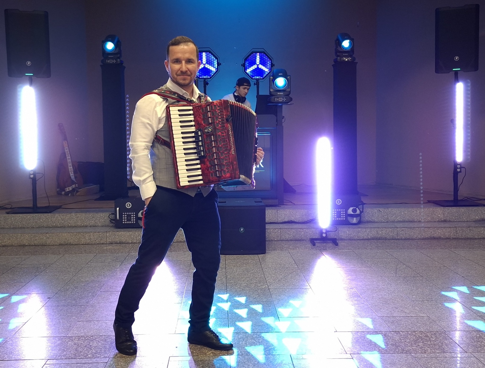 Akordeonista w szarej kamizelce, na scenie z oświetleniem, drugi muzyk w tle. Występ na żywo, akordeon w kolorze czerwonym.
