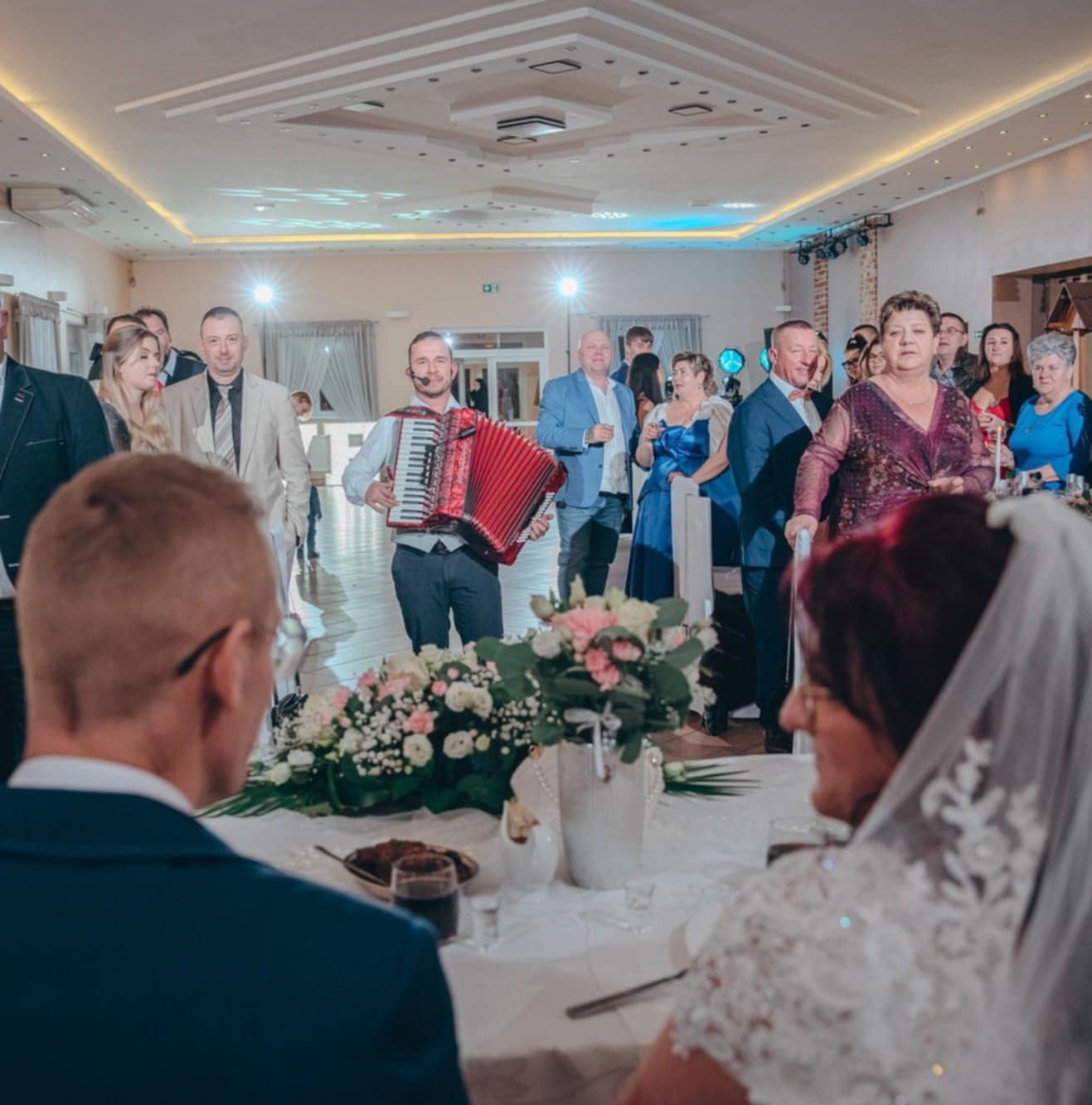 Akordeonista z mikrofonem gra dla gości weselnych w sali bankietowej, widok zza stołu pary młodej. Dekoracje kwiatowe i eleganckie oświetlenie.