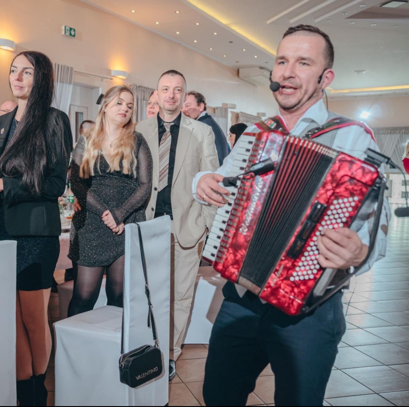 Akordeonista z mikrofonem gra na czerwonym akordeonie na tle gości weselnych w jasnej sali. Widoczny mężczyzna w beżowym garniturze i kobiety w sukienkach.