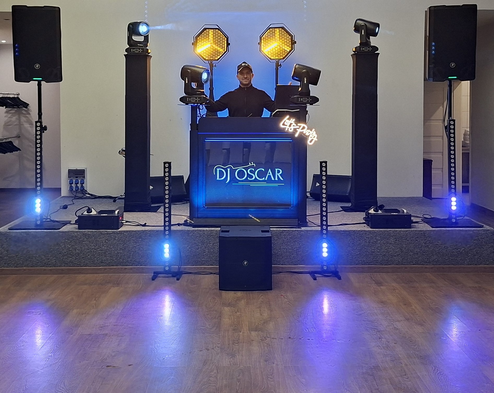 DJ Oscar za konsoletą z neonowym napisem 'Let's Party', otoczony sprzętem nagłośnieniowym i oświetleniem scenicznym. Scena oświetlona niebieskim światłem.