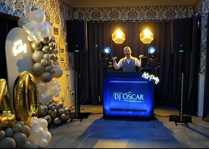 DJ Oscar na stanowisku z oświetleniem i nagłośnieniem, dekoracja balonowa w odcieniach bieli i złota. DJ w szarej kamizelce, uśmiechnięty, gotowy do występu.