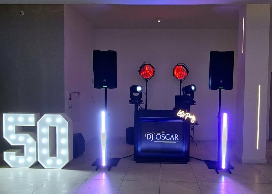 Oświetlona dekoracja '50' obok stanowiska DJ'a z logo 'DJ Oscar', głośnikami i reflektorami na tle jasnej ściany. Scena gotowa na imprezę.