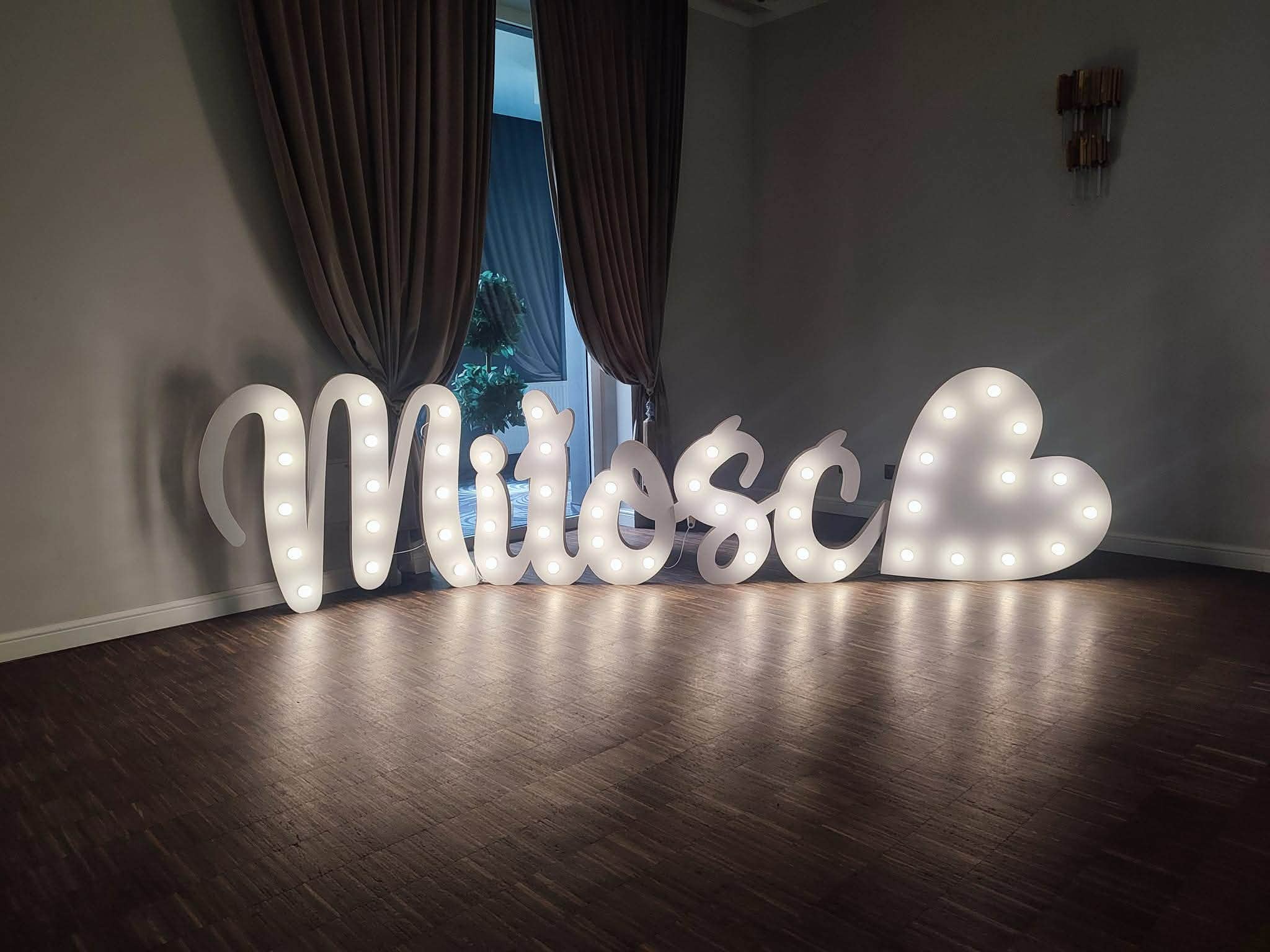 Świecący napis 'Miłość' z żarówkami na drewnianej podłodze, ustawiony przed oknem z zasłonami. Dekoracja na imprezę, romantyczny nastrój.