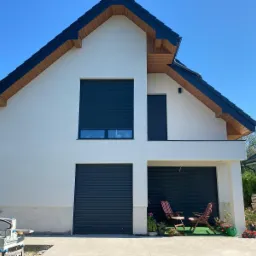 Nowoczesny dom jednorodzinny z białymi ścianami, ciemną stolarką okienną i bramami garażowymi z roletami zewnętrznymi. Na podjeździe dwa krzesła ogrodowe i rośliny w donicach.