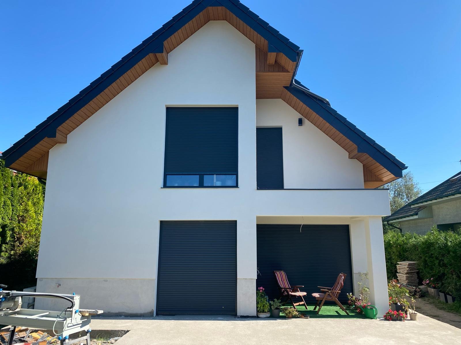 Nowoczesny dom jednorodzinny z białymi ścianami, ciemną stolarką okienną i bramami garażowymi z roletami zewnętrznymi. Na podjeździe dwa krzesła ogrodowe i rośliny w donicach.