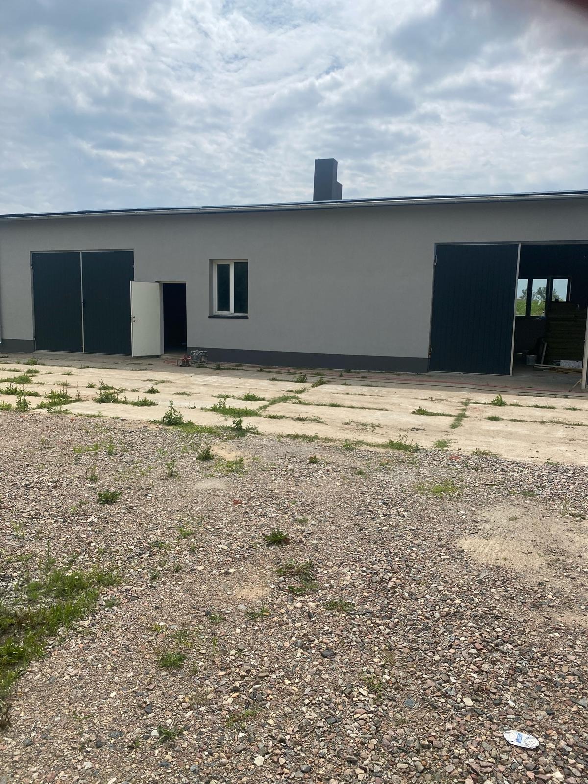 Szary budynek gospodarczy z dwoma bramami garażowymi, jedną otwartą, na żwirowym terenie z betonowymi płytami i rosnącą trawą.