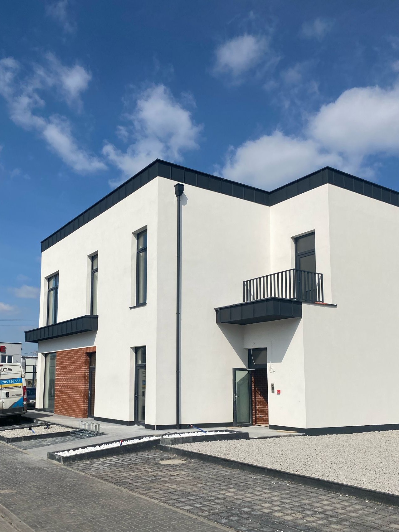 Nowoczesny, dwukondygnacyjny dom z białym tynkiem i fragmentem elewacji z cegły, czarne obramowania okien, balkon z metalową barierką, widoczne elementy zagospodarowania terenu.