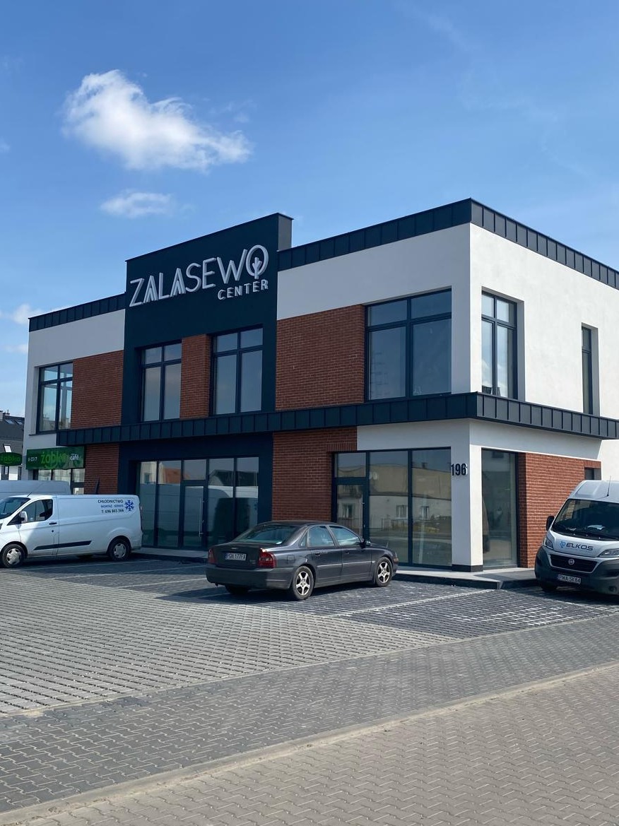 Dwukondygnacyjny budynek biurowy z białymi ścianami, elementami z cegły i ciemnymi akcentami, z napisem 'Zalasewo Center' nad wejściem, widok z zewnątrz na parking z dwoma samochodami dostawczymi...