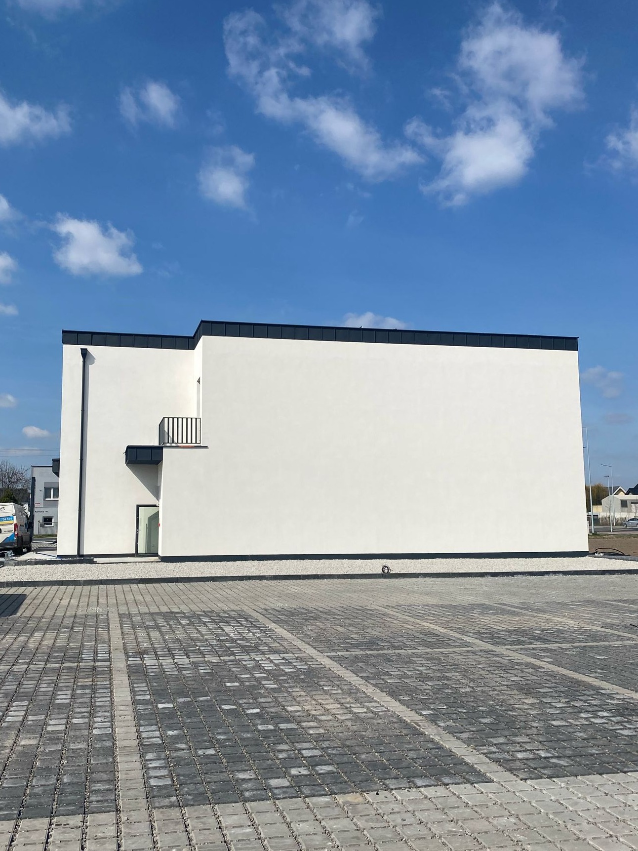 Nowoczesny, biały dom murowany z płaskim dachem i małym balkonem, widok z poziomu parkingu z kostki brukowej, błękitne niebo z chmurami w tle.