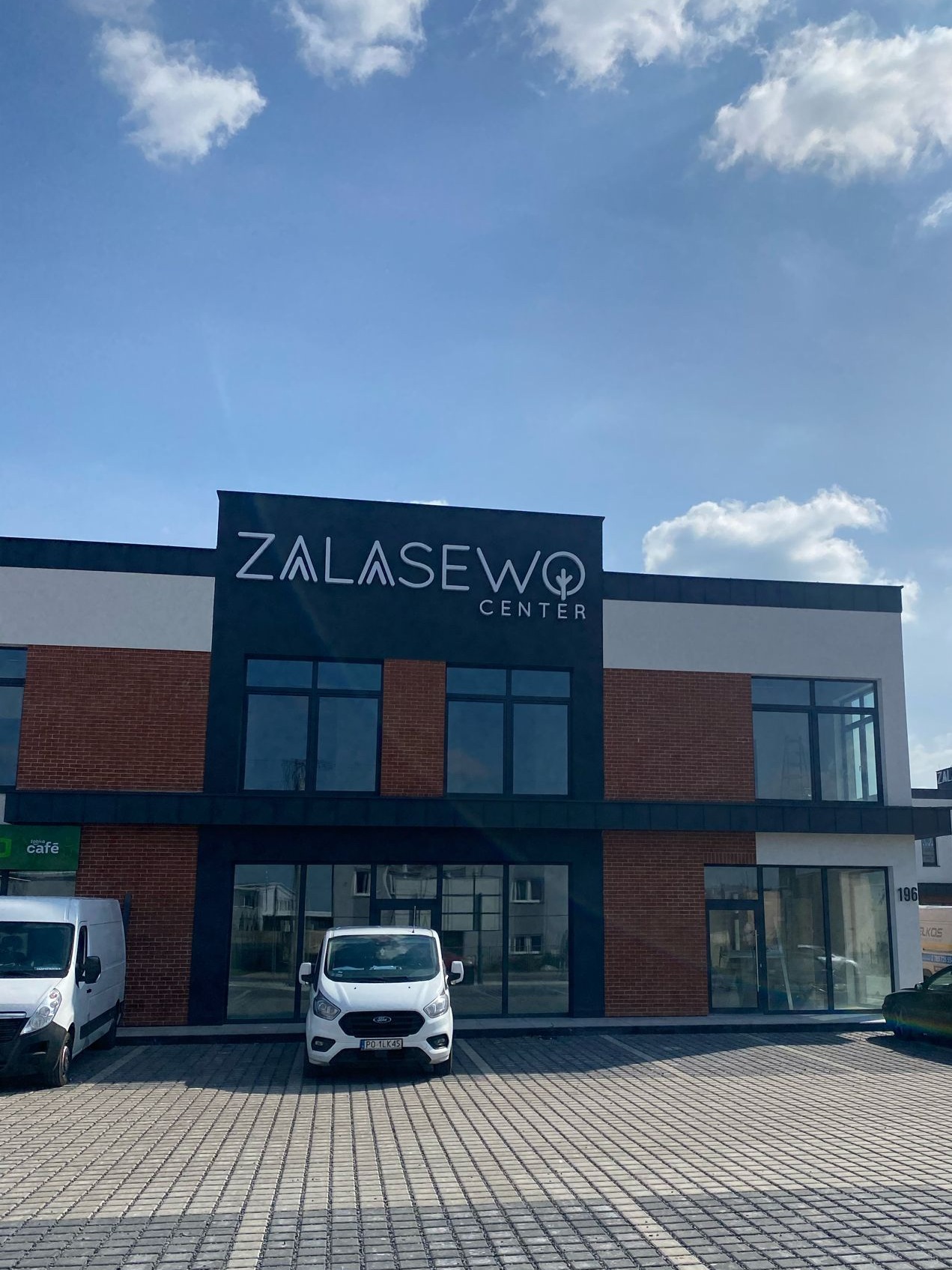 Dwukondygnacyjny budynek biurowy z ceglaną elewacją i dużymi oknami, z napisem 'Zalasewo Center' na górze i zaparkowanymi samochodami dostawczymi na brukowanym parkingu przed nim.