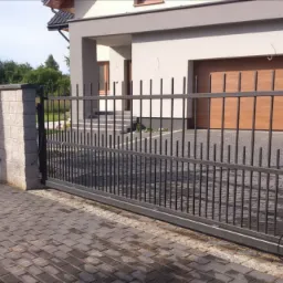 Przesuwna brama wjazdowa z metalowymi prętami, osadzona między murowanymi słupkami, widok z zewnątrz na posesję z kostki brukowej i nowoczesny dom z garażem.