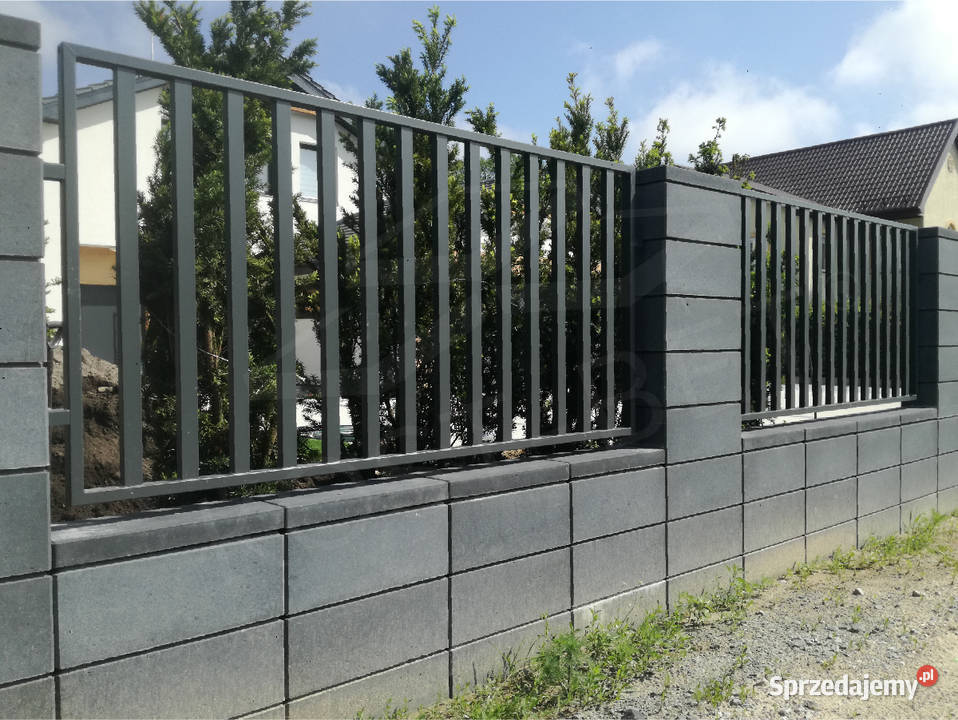 Szary, modułowy mur oporowy z metalowymi przęsłami tworzący ogrodzenie posesji, widok z zewnątrz na tle zieleni i zabudowań.